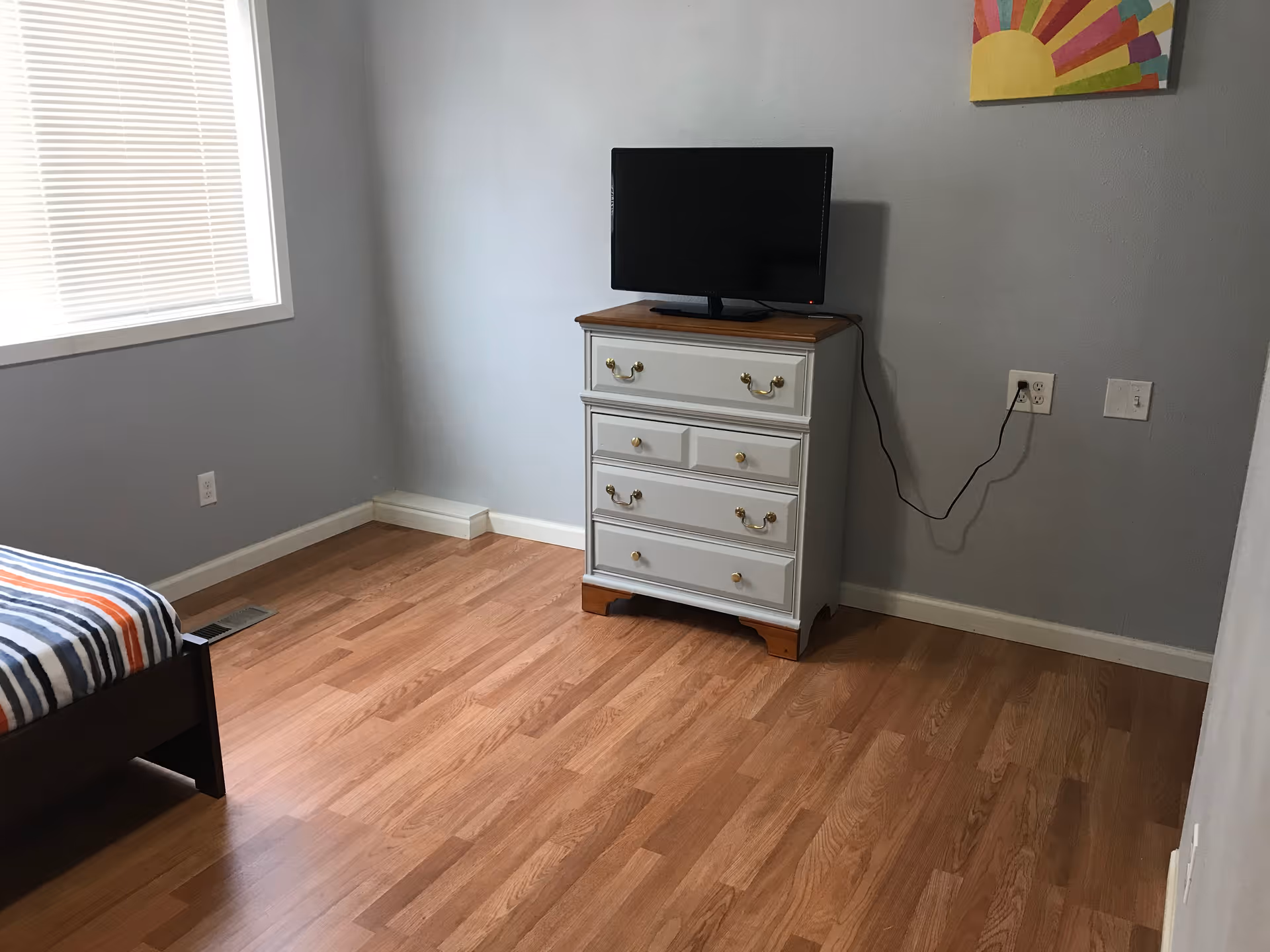 A bedroom with light gray walls and wood flooring. There is a window with closed blinds on the left wall. A bed with a striped blanket is partially visible on the left side. Against the far wall, there is a white dresser with brass handles and a wooden top, on which a flat-screen TV is placed. A colorful abstract painting is hung on the wall above the dresser. An electrical outlet with a plugged-in TV power cord is visible on the right wall.