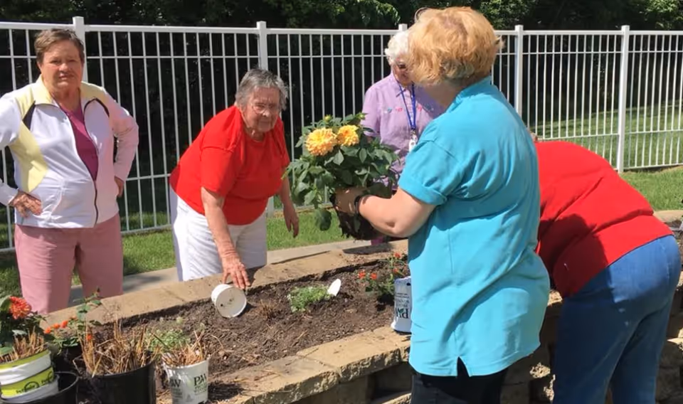 A group of elderly women gardening together outdoors by a raised stone planter bed with flowers and plants, surrounded by a white metal fence and green grass.
