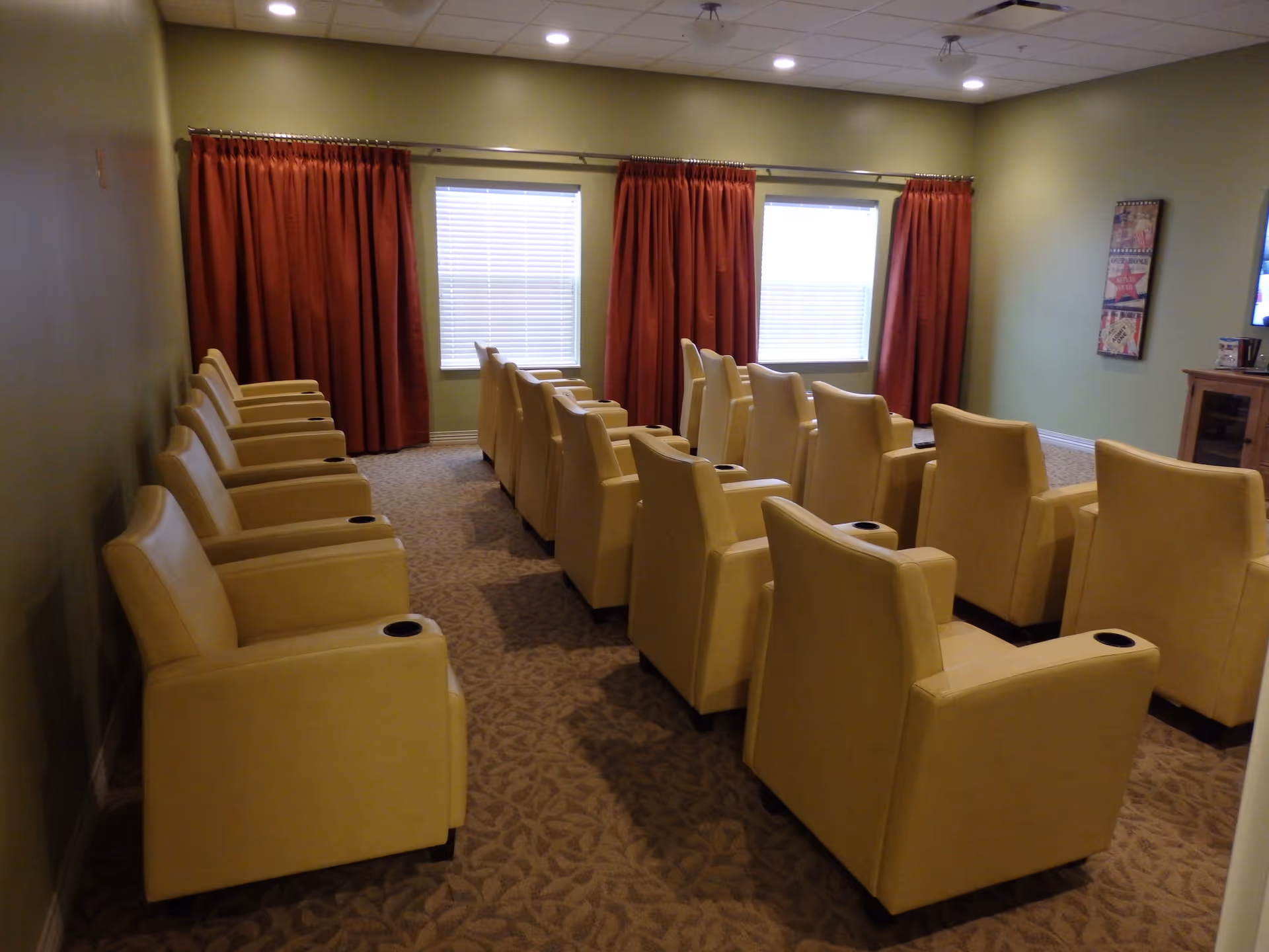 A small theater room with three rows of beige recliner chairs facing a TV mounted on the wall. The room has green walls, two windows with red curtains, and carpeted floor with a leaf pattern.