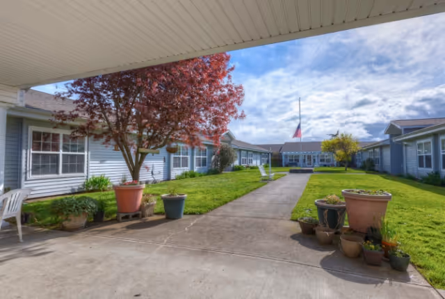 Outdoor courtyard area of a senior living facility with a concrete walkway, green grass, potted plants, and a tree with red leaves. Single-story buildings with blue siding and white trim surround the courtyard under a partly cloudy sky.