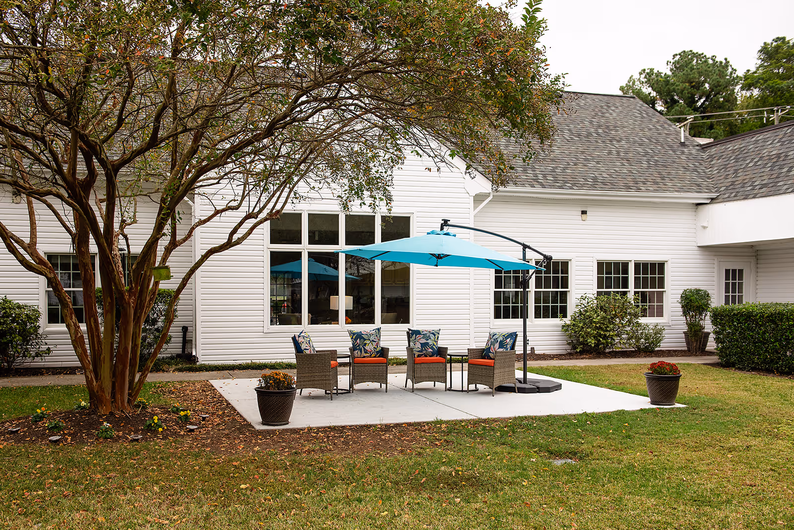 Patio with four wicker chairs and a teal umbrella on a concrete pad in a grassy courtyard beside a white building.