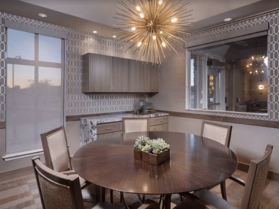 A modern dining room with a round wooden table surrounded by six upholstered chairs. A decorative centerpiece with small plants is on the table. The room features patterned wallpaper, a large window with a roller shade, wooden cabinets, and a unique starburst chandelier hanging from the ceiling. A large mirror on one wall reflects part of the room and chandelier.