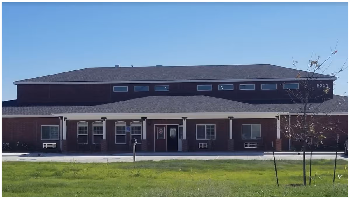 Front exterior view of a single-story brick building with a dark shingled roof, several windows, and a covered entrance. There is a small tree and green grass in the foreground under a clear blue sky.