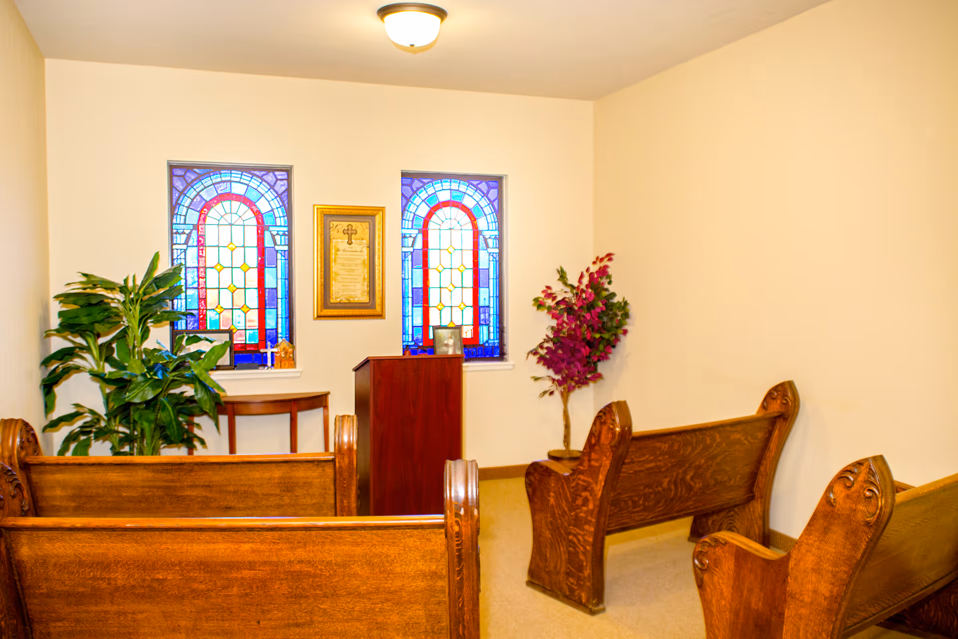 Small chapel room with wooden pews, two stained glass windows, a wooden podium, framed religious text on the wall, and potted plants in the corners.