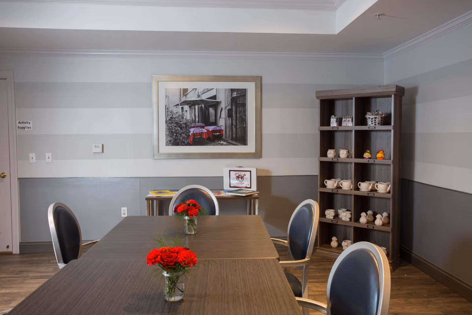 Activity/dining room with a long table and chairs, vases of red flowers, shelving with cups and crafts, and framed artwork on the wall.