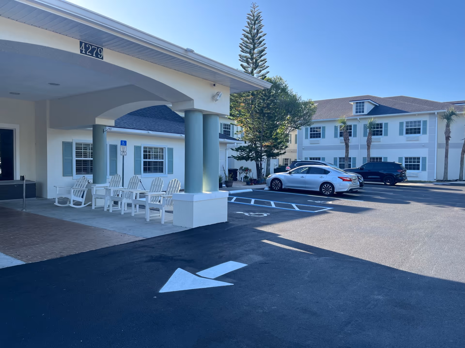 Exterior view of Cedar Creek Assisted Living facility showing the entrance with white rocking chairs on the porch, a covered area with columns, a parking lot with cars, and a clear blue sky.