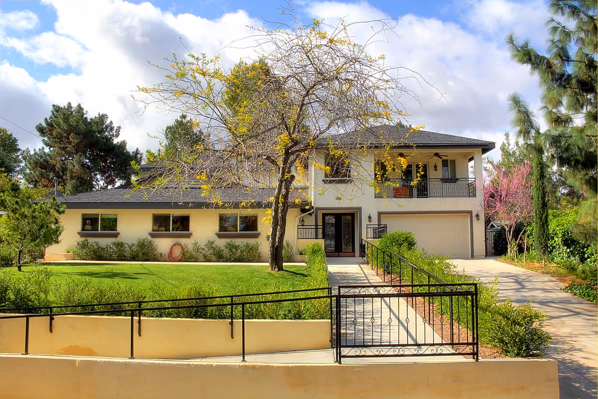 Front exterior of a two-story home with a driveway, garage, balcony, fenced walkway, and a lawn with a tree.