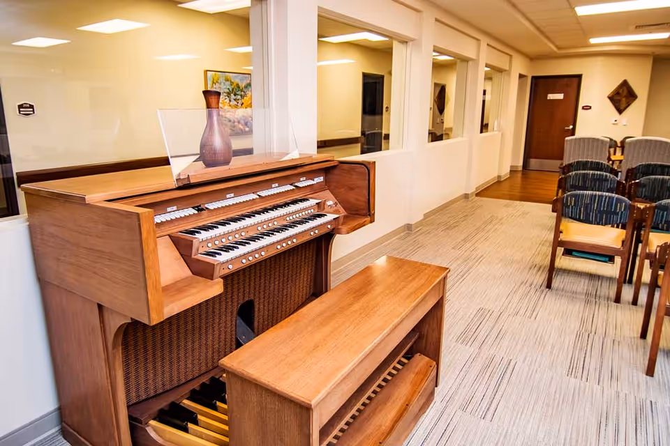 Interior view of a senior living facility hallway featuring a wooden organ with a bench in the foreground. The hallway has light-colored walls, carpeted flooring, and several chairs arranged in rows along the right side. There are windows along the left wall and a closed door at the end of the hallway.