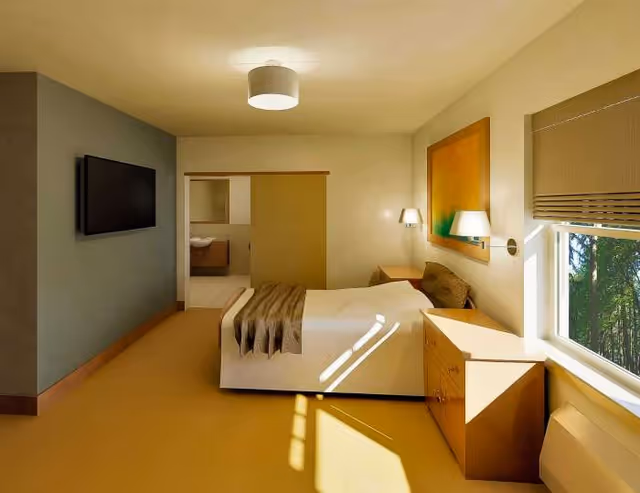 A tidy hotel-style bedroom with a bed, dresser, TV, window, and bathroom doorway.