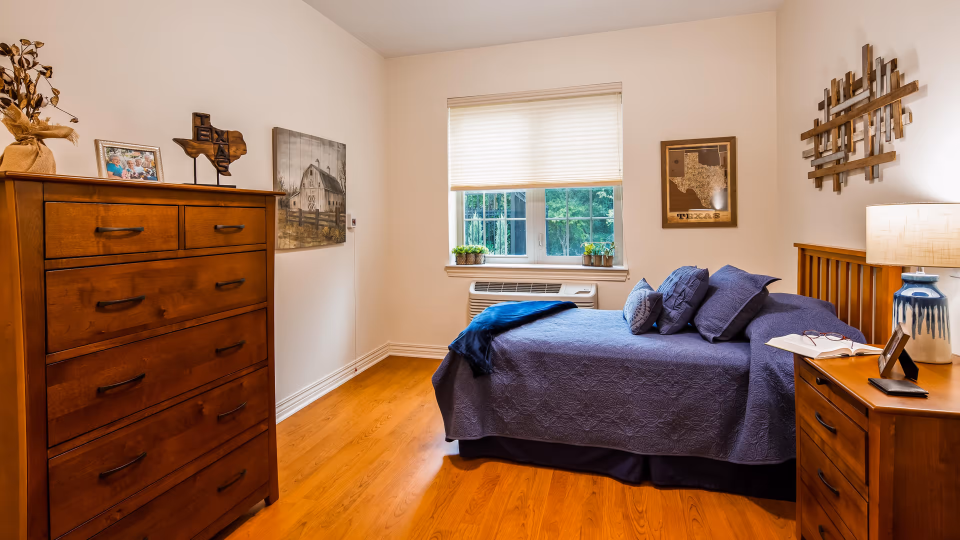 A cozy bedroom with wooden furniture including a tall dresser and a nightstand with a lamp. The bed is made with a blue quilt and several pillows. There are framed pictures and decorative items on the walls, and a window with a white blind lets in natural light. The floor is hardwood.