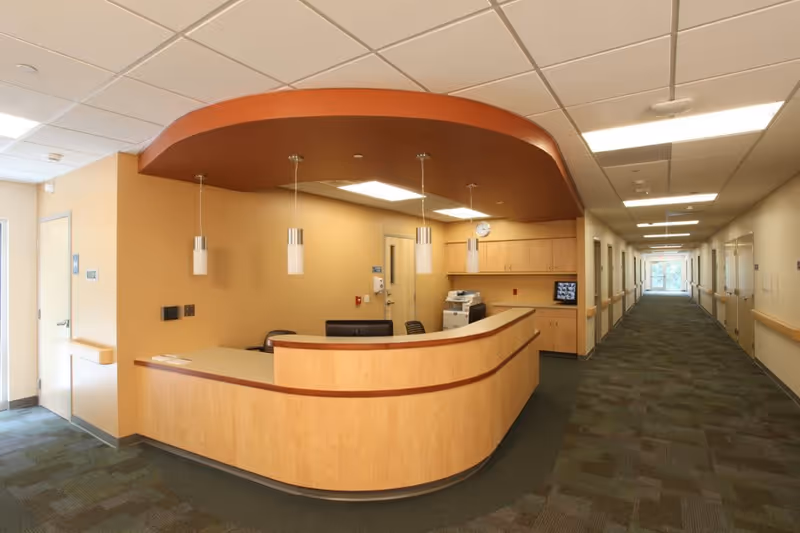 Reception desk area inside a senior living facility with a curved wooden counter, three pendant lights hanging from the ceiling, and a long hallway with multiple doors and handrails on both sides.