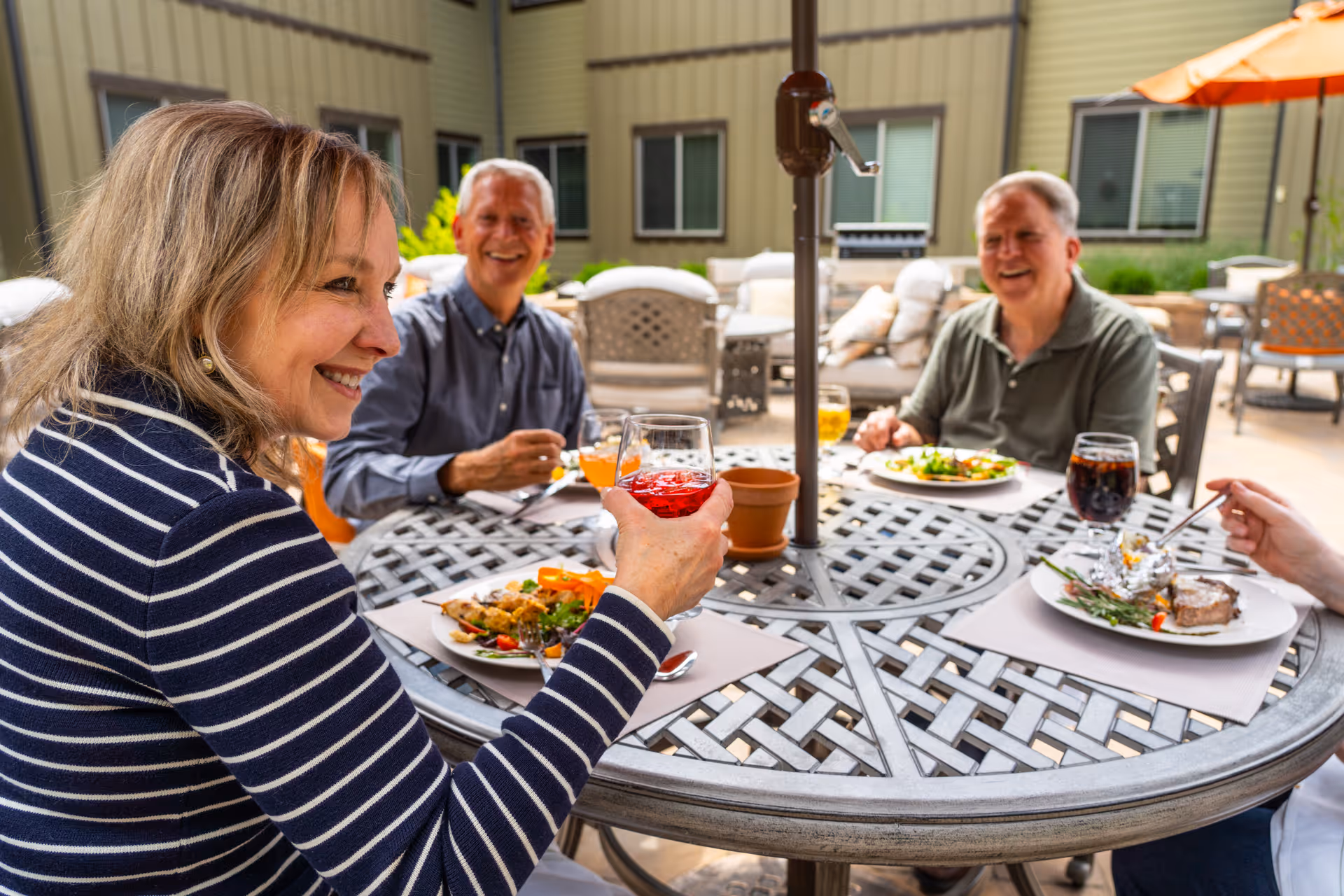 Three elderly people sitting around a round outdoor table enjoying a meal and drinks together on a patio with cushioned chairs and umbrellas in the background.