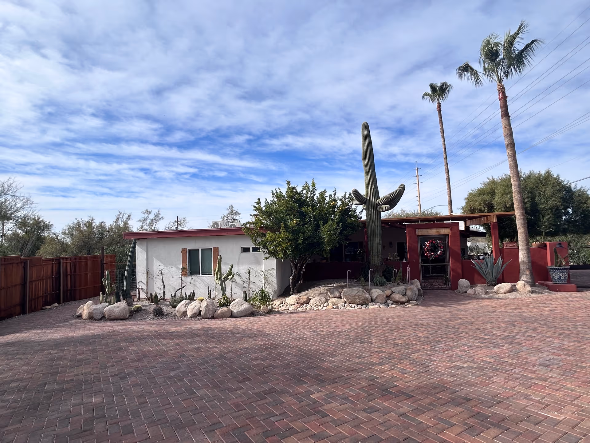 Exterior view of Casa de Abuelos at Edgewood showing a single-story building with white and red walls, a large cactus, palm trees, and a paved brick driveway under a partly cloudy sky.