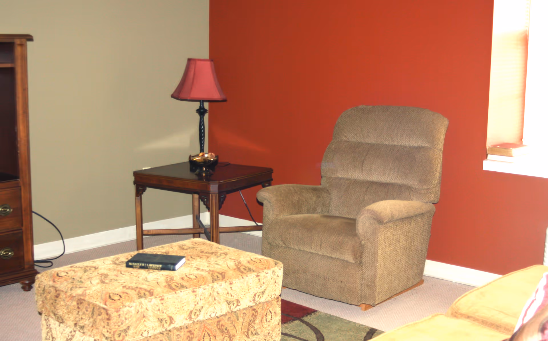 A cozy living room corner with a brown upholstered recliner chair, a wooden side table with a red lampshade, a patterned ottoman with a book on top, and part of a wooden cabinet. The walls are painted in two tones: beige and burnt orange, with a window letting in natural light.