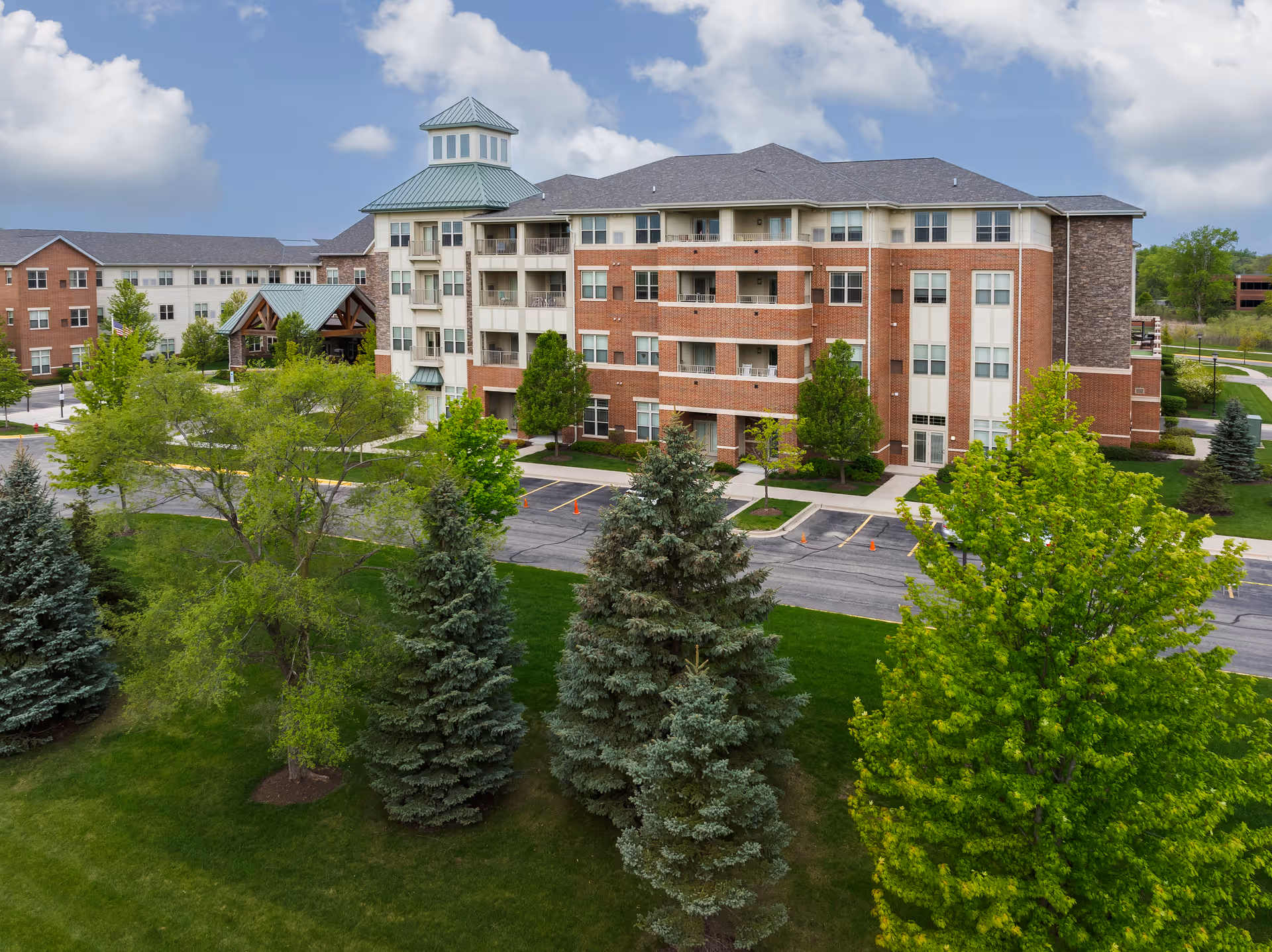 Exterior view of a multi-story senior living facility building with a mix of brick and light-colored siding, surrounded by green trees and a well-maintained lawn under a partly cloudy sky.