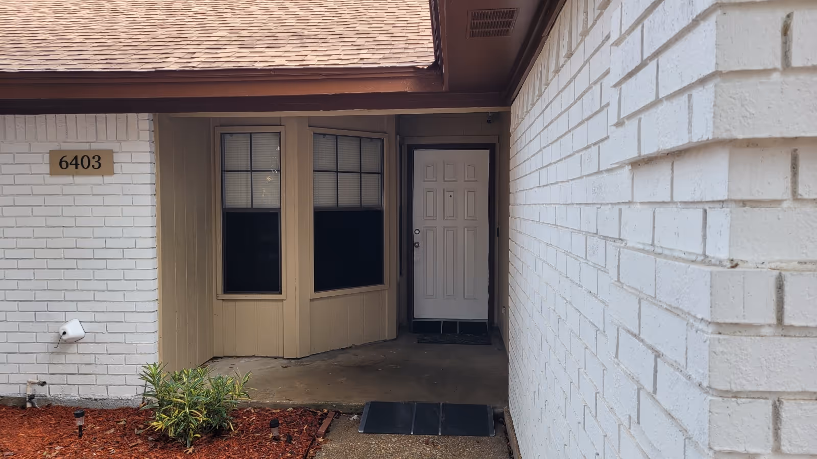 Front entrance of a white-brick building with a recessed doorway, bay window, house number 6403, and a small ramp.