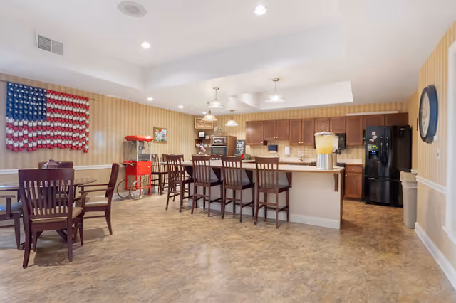 A spacious dining area with wooden tables and chairs, a kitchen island with bar stools, a popcorn machine, and a large American flag decoration on the wall. The kitchen area has wooden cabinets, a black refrigerator, and a large beverage dispenser on the counter.