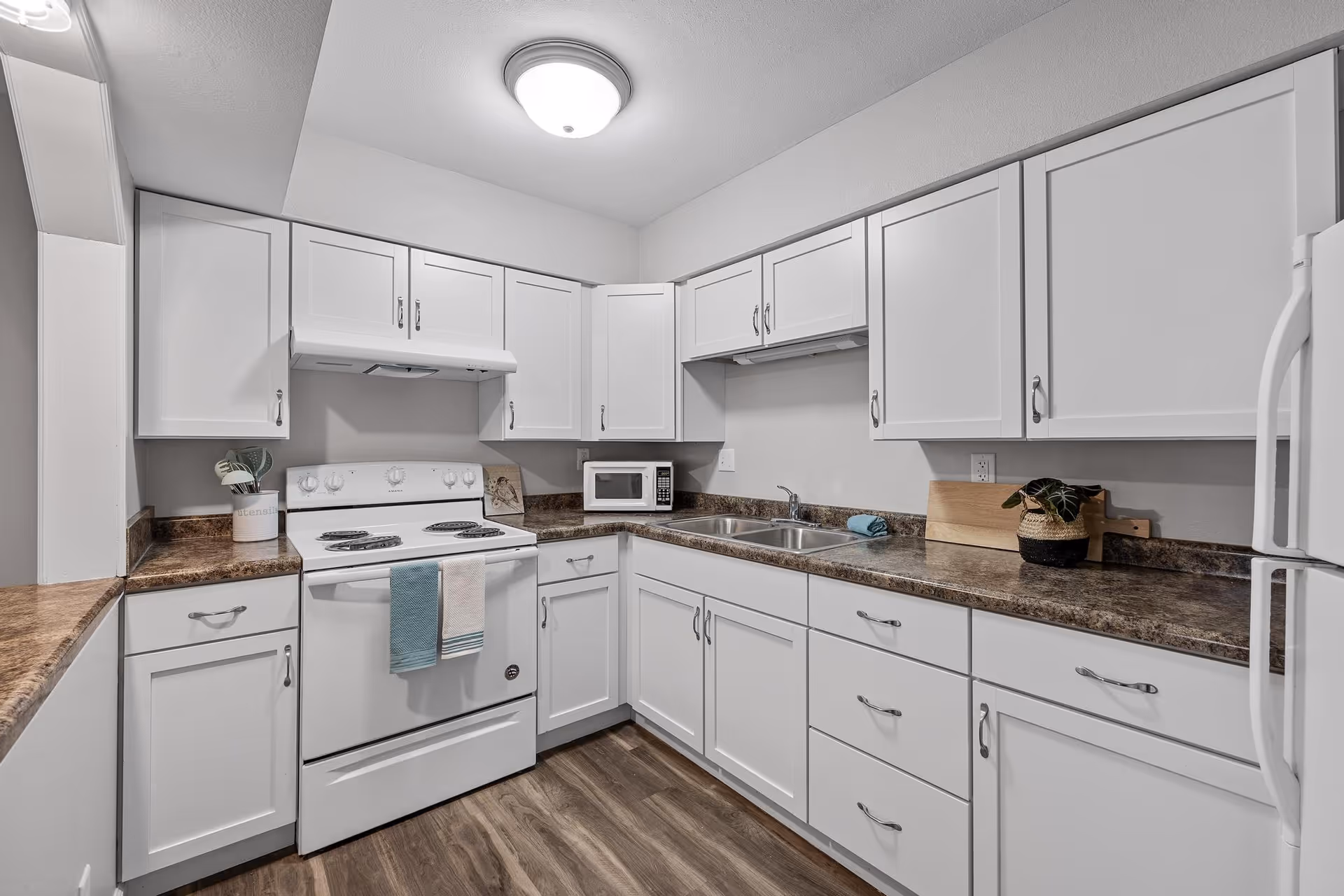 A clean and modern kitchen with white cabinets, a white electric stove with two towels hanging on the handle, a microwave on the countertop, a double sink, and a small potted plant on the right side. The countertops are brown with a marble pattern, and the floor has a wood finish. The kitchen is well-lit with a ceiling light.