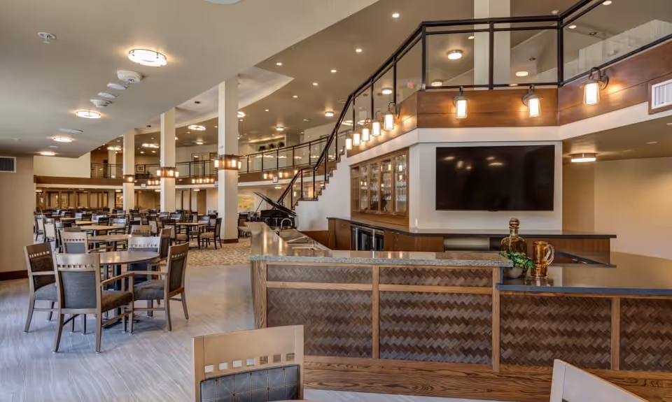 Spacious dining area with multiple wooden tables and chairs arranged neatly. A large counter with a granite top is in the foreground, featuring decorative items and a plant. The room has a high ceiling with recessed lighting and a second-floor balcony with glass railings. A large flat-screen TV is mounted on the wall behind the counter. The space is well-lit and has a modern, welcoming atmosphere.