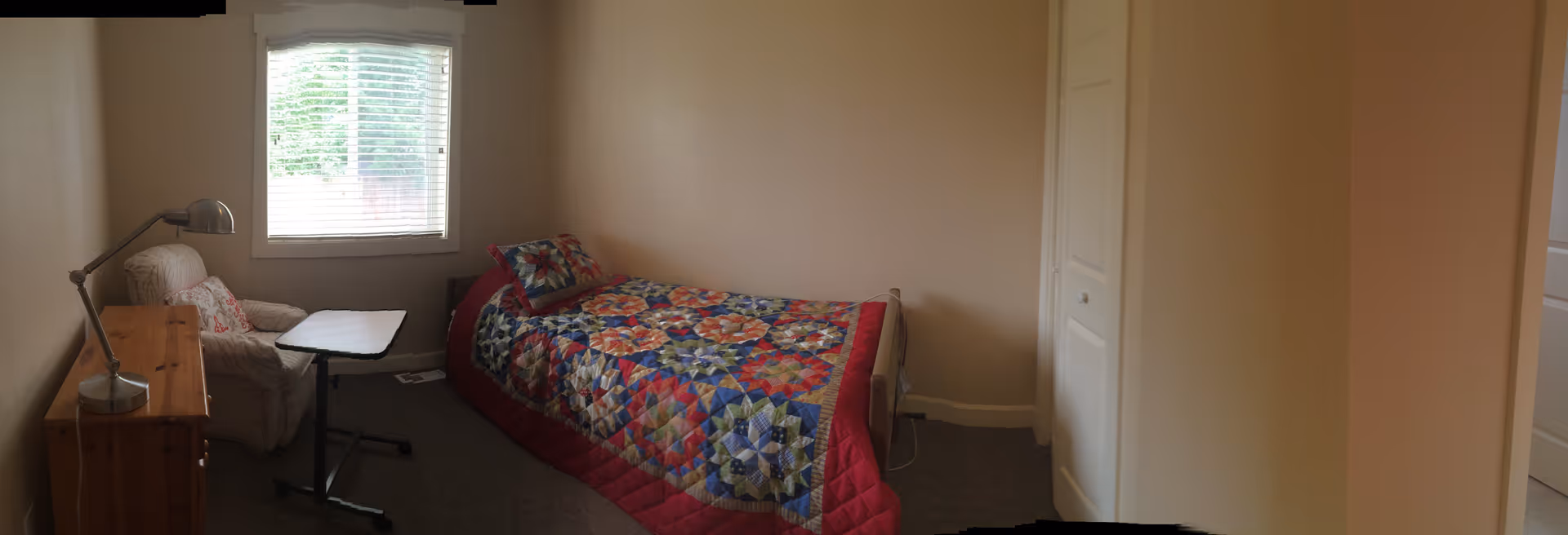 A small bedroom with a single bed covered with a colorful patchwork quilt. Next to the bed is a white armchair with a floral pillow and a small adjustable table. A wooden dresser with a desk lamp is positioned against the opposite wall. A window with white blinds lets in natural light.