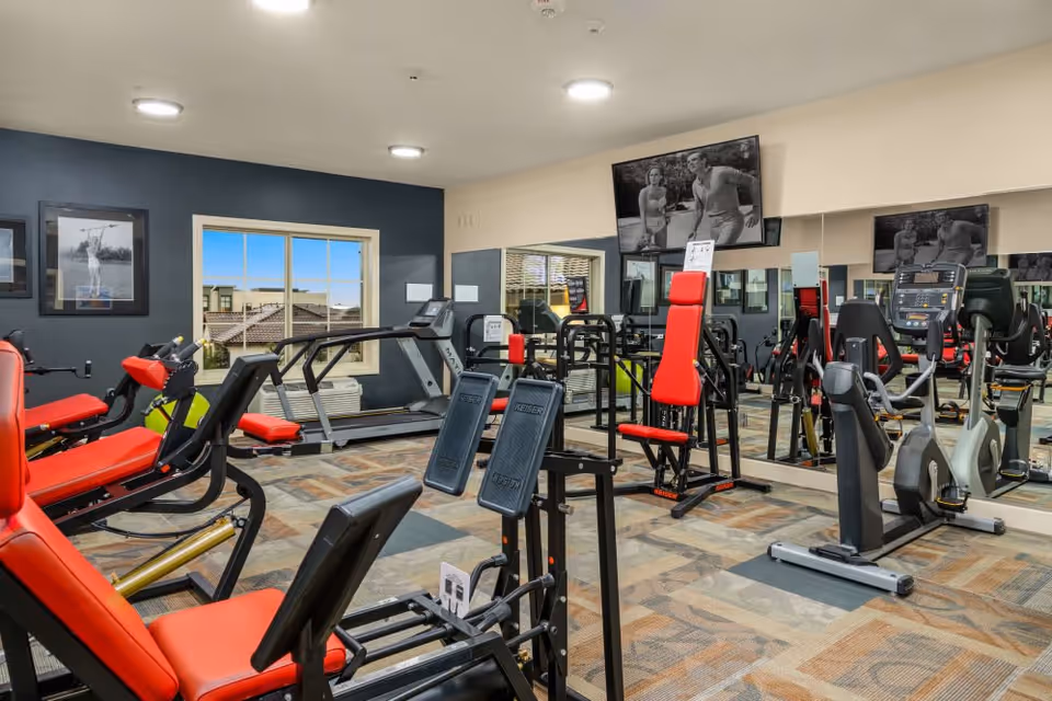 Fitness room with cardio and strength machines featuring red padded seats, wall mirrors, and mounted TVs.