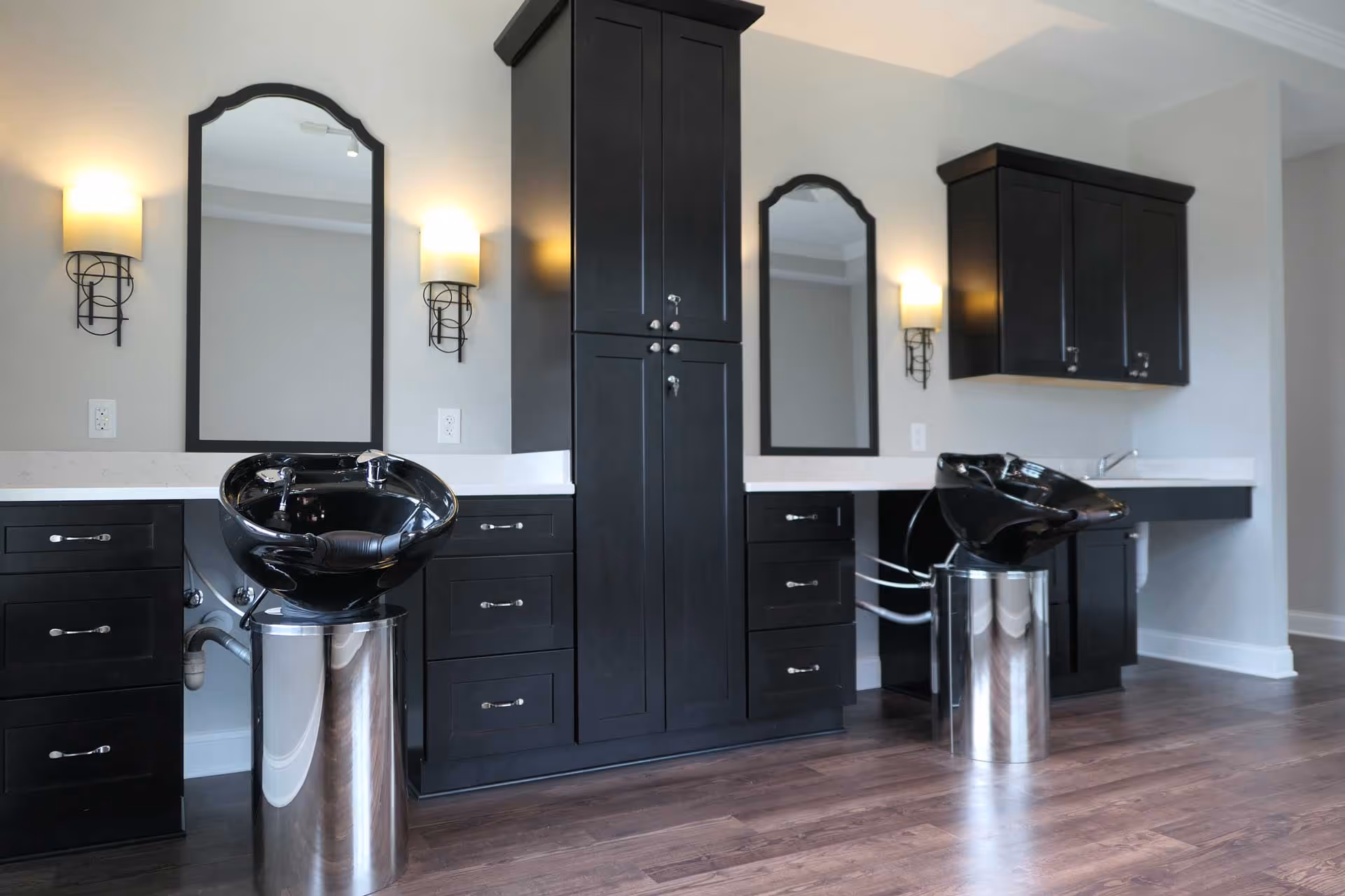 Interior salon area with two black shampoo basins, dark cabinetry, mirrors, and wall sconces.