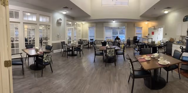 A spacious dining room in a senior living facility with multiple tables and chairs arranged neatly. The room has large windows allowing natural light, light-colored walls, and a wood-style floor. Some tables are set with cups and plates, and there is a person sitting alone near the windows at the far end of the room. The room also features a small kitchenette area with cabinets and appliances on the right side.