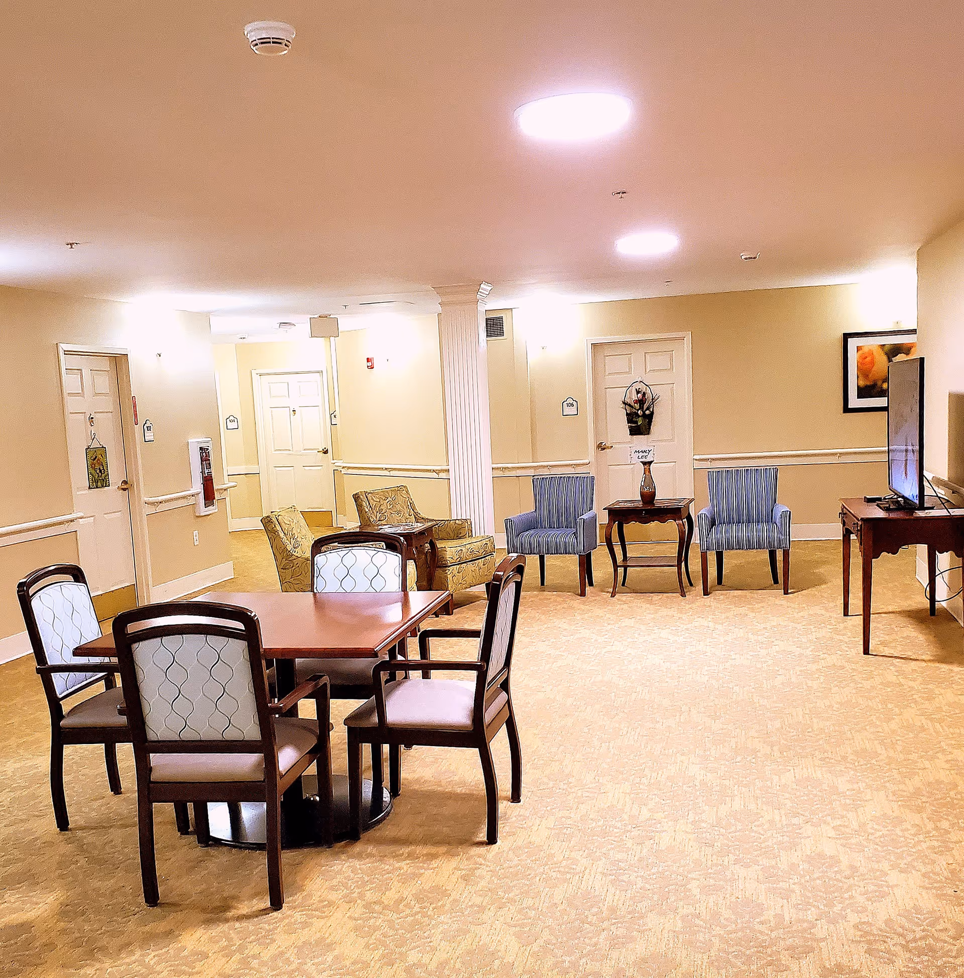 A well-lit senior living facility common area with a square wooden table surrounded by four cushioned chairs in the foreground. In the background, there are two patterned armchairs and two blue striped chairs with a small wooden table between them holding a decorative vase. A flat-screen TV is mounted on a wooden stand to the right. The walls are beige with white trim, and several doors and framed artwork are visible.