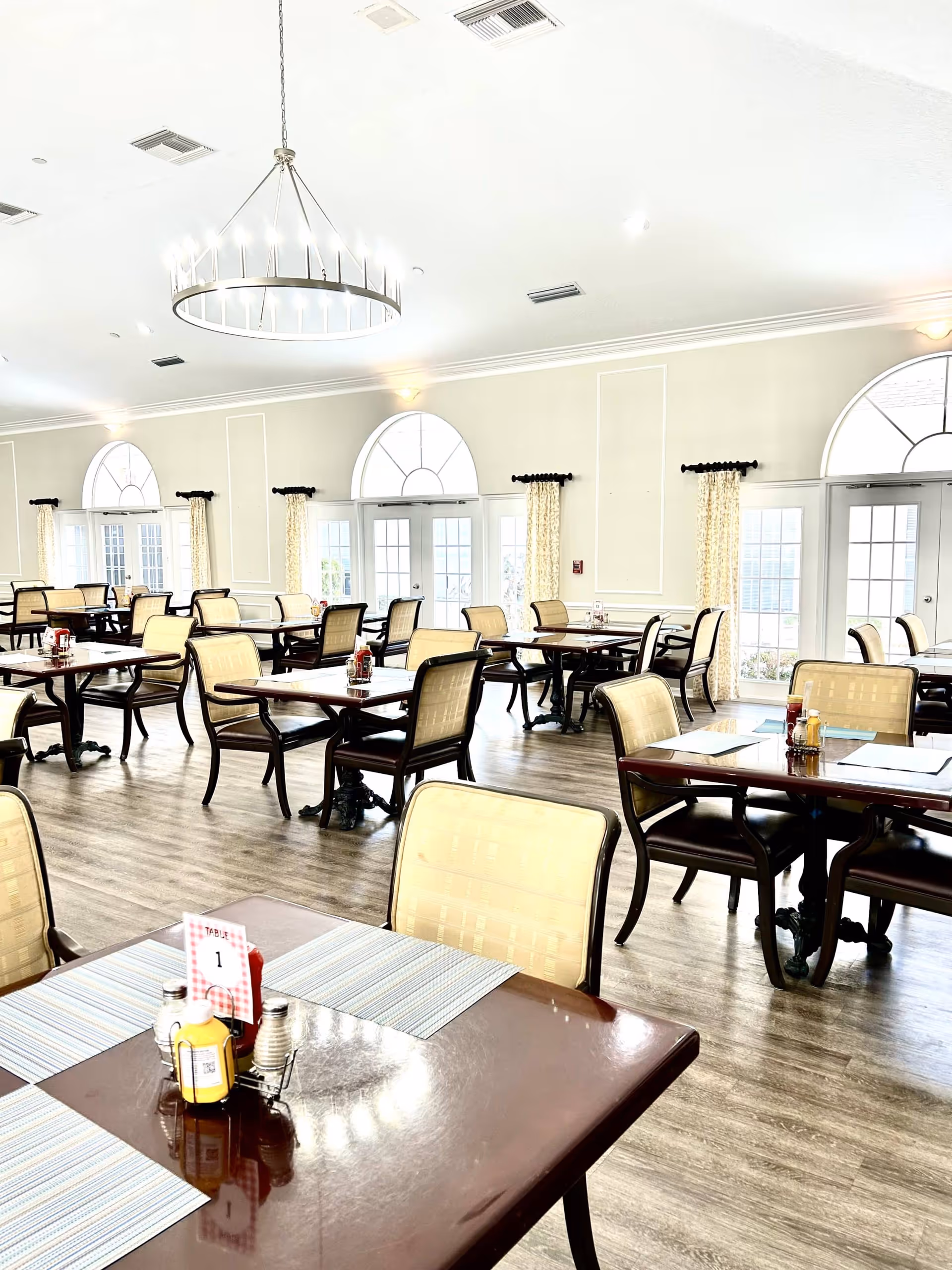 Bright and spacious dining room with multiple wooden tables and cushioned chairs arranged neatly. Each table has placemats and condiments including ketchup, mustard, salt, and pepper. Large arched windows with light curtains allow natural light to fill the room. A modern circular chandelier hangs from the ceiling.