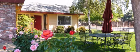 Single-story house with a well-kept lawn, rose garden, and metal patio table under an umbrella.