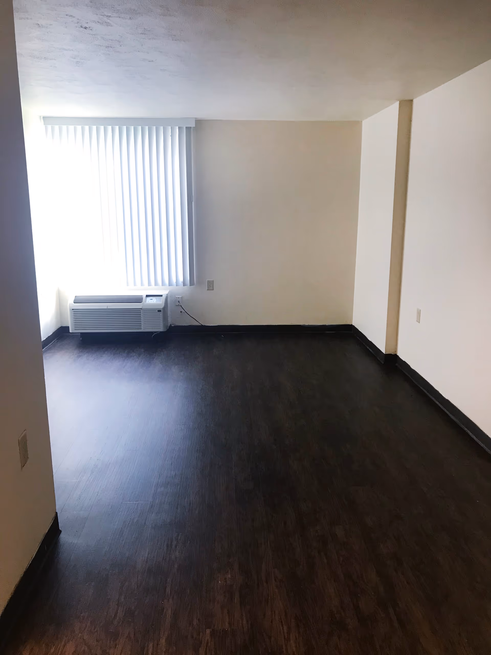 Empty room with dark wood flooring, beige walls, a window with vertical blinds, and a wall-mounted air conditioning unit beneath the window.