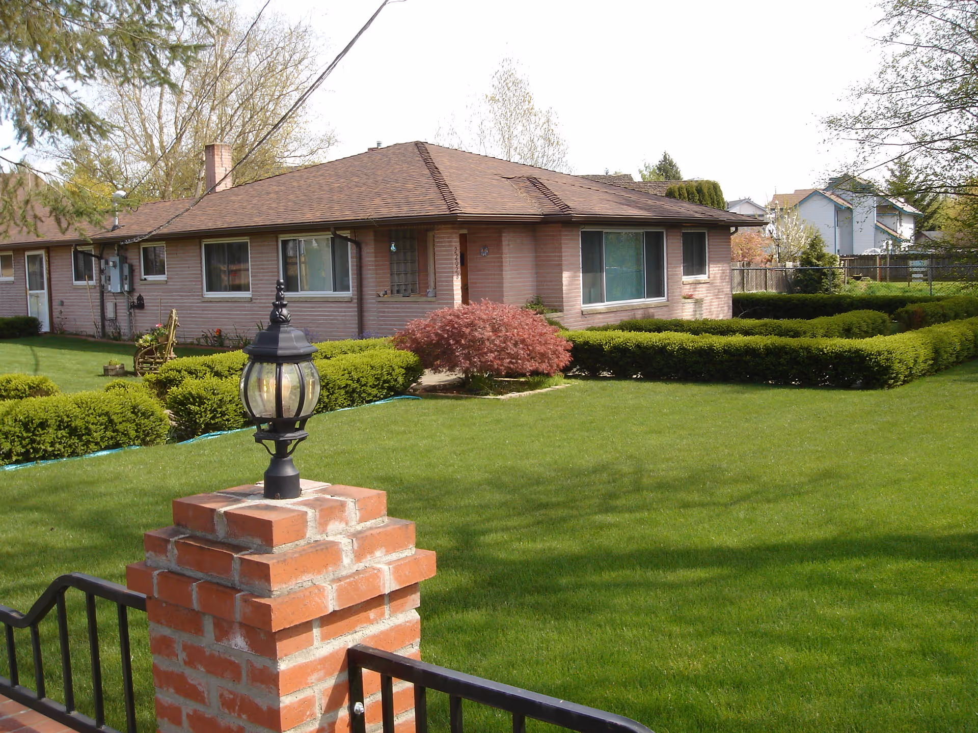 A single-story brick building with a brown roof surrounded by a well-maintained green lawn and neatly trimmed hedges. There is a brick pillar with a black lantern on top in the foreground, and trees and other houses are visible in the background.