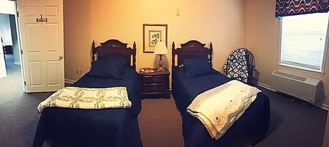 A bedroom with two twin beds, each with dark blue bedding and a folded blanket at the foot. Between the beds is a wooden nightstand with a lamp and a framed picture on the wall above it. There is a window with a patterned valance on the right side and a chair with a blanket draped over it next to the window. A door is open on the left side of the room.