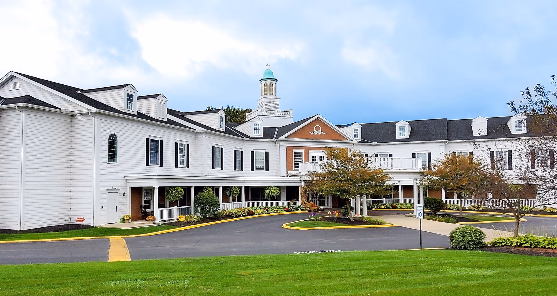 Exterior view of a large, white, multi-story assisted living facility building with black shutters and a cupola on the roof. The building is surrounded by a well-maintained lawn, trees, and a paved driveway with a roundabout in front.