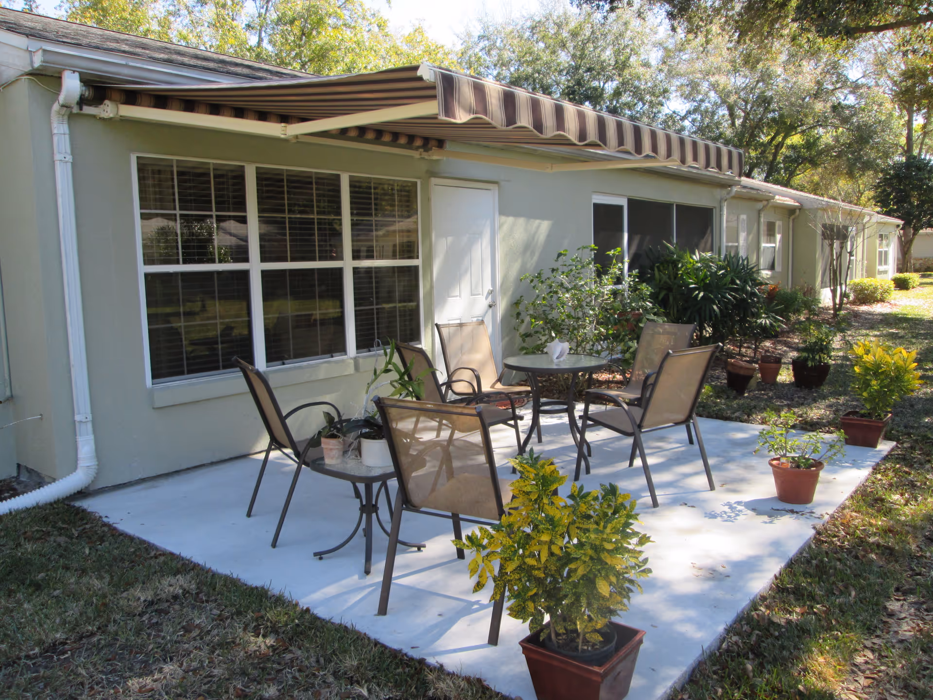 Outdoor patio area with a concrete floor, featuring a round glass table surrounded by four mesh chairs. Several potted plants are placed around the patio. The patio is attached to a light-colored building with large windows and a white door. A striped retractable awning extends over the patio, and trees and shrubs are visible in the background.
