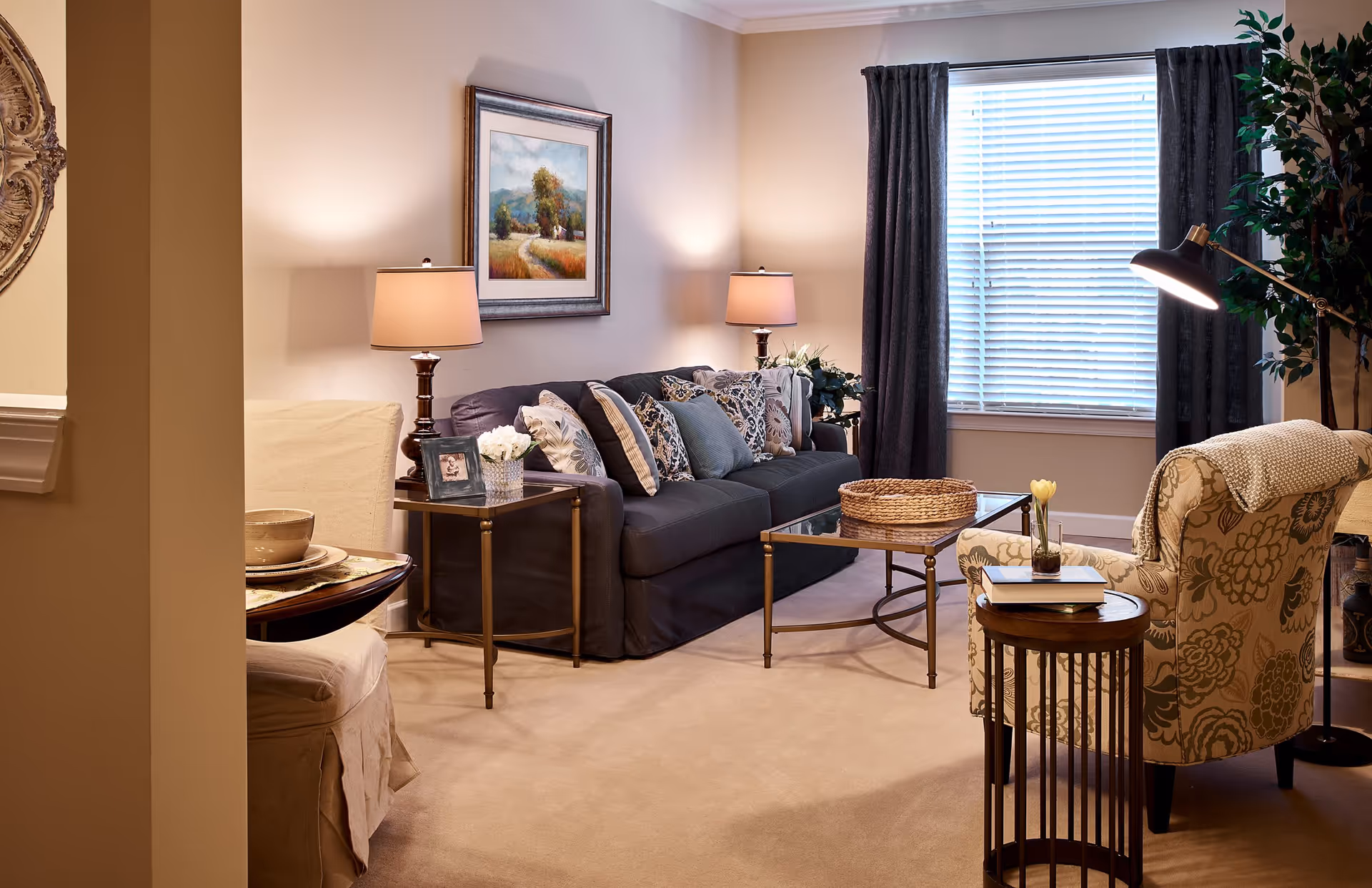 A cozy living room with a dark gray sofa adorned with multiple patterned pillows, two side tables with lamps, a glass coffee table with a woven basket, a patterned armchair with a throw blanket, a small round wooden side table with books and a flower vase, a large window with blinds and dark curtains, and a potted plant in the corner.