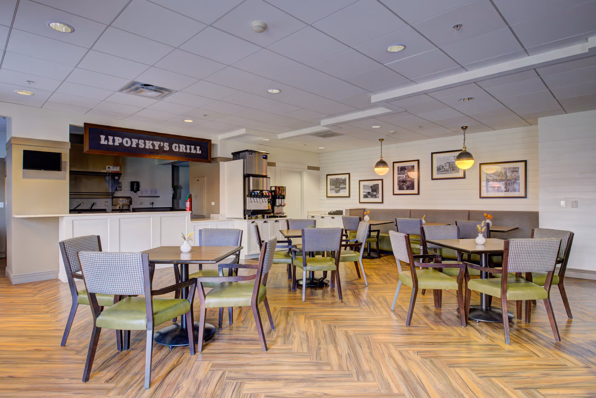 A dining area with several tables and chairs arranged on a wood-patterned floor. The chairs have green seats and patterned backs. On the left side, there is a counter with a sign above it reading 'Lipofsky's Grill'. Behind the counter, kitchen equipment and a soda fountain machine are visible. The walls are decorated with framed pictures and two hanging pendant lights.