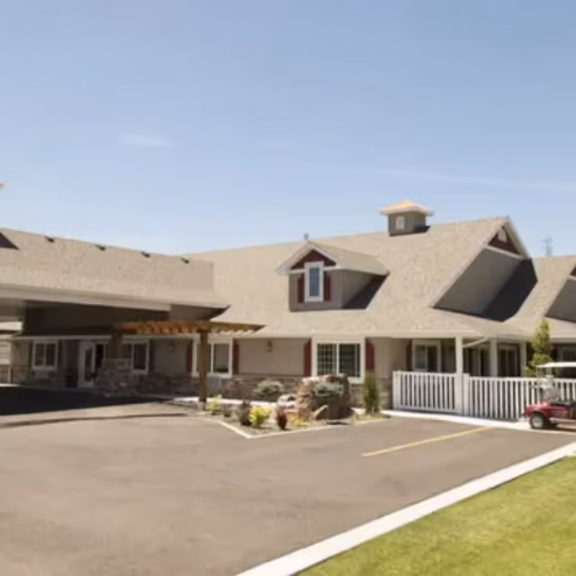 Front exterior of a single-story memory care building featuring a covered entrance, dormer windows, landscaped planters, and a small parking area.