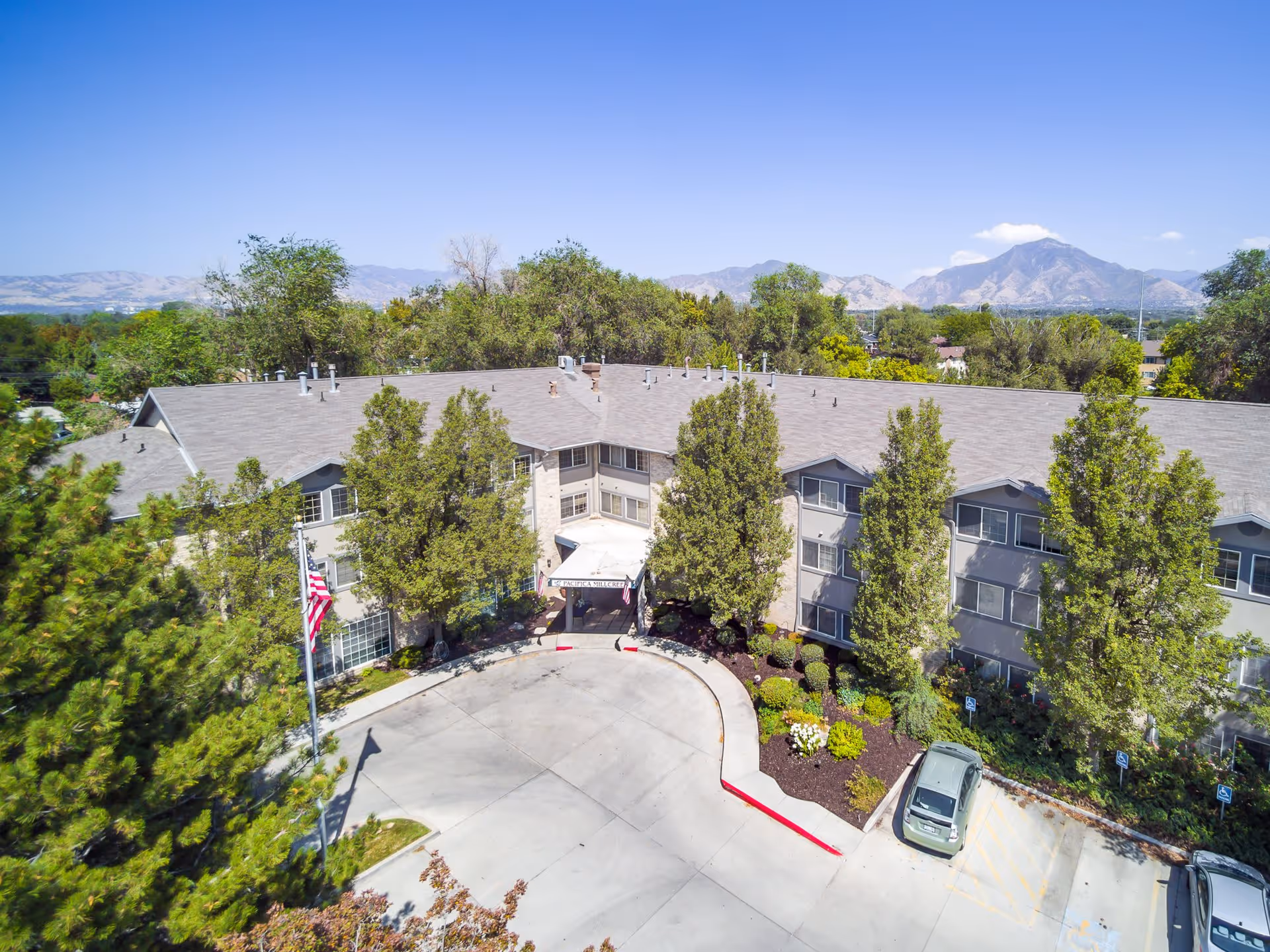 Aerial view of a three-story senior living facility building with a gray roof and beige exterior, surrounded by trees and landscaping. There is a circular driveway at the entrance with an American flag on a flagpole. Mountains are visible in the background under a clear blue sky.