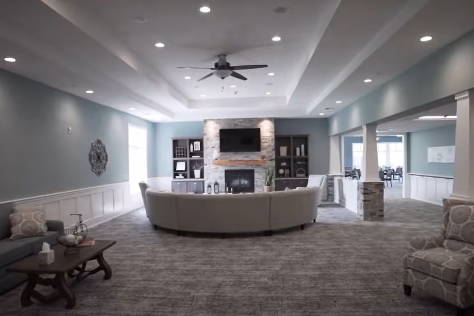 A spacious living room area with a curved beige sofa facing a stone fireplace with a mounted flat-screen TV above it. The room has light blue walls, recessed ceiling lights, a ceiling fan, and built-in dark wood shelves flanking the fireplace. There are additional seating areas with patterned armchairs and a coffee table with decorative items. The room opens into a dining area with tables and chairs visible in the background.