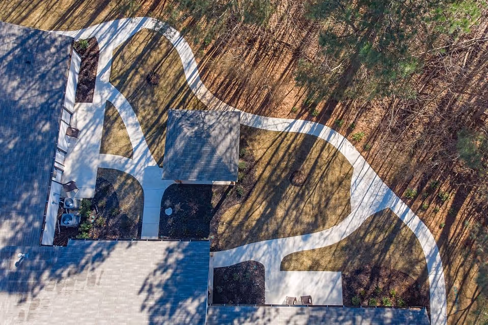 Aerial view of a senior living facility's outdoor garden area with winding concrete pathways, a small covered pavilion, landscaped garden beds, and surrounding trees casting long shadows.