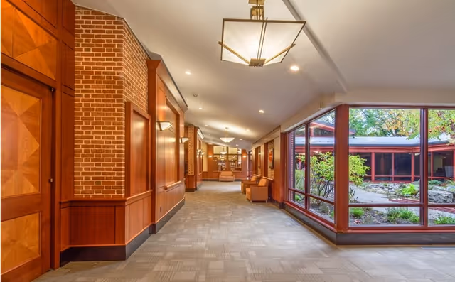 A well-lit hallway with carpeted flooring, wood-paneled walls, and brick accents. Large windows on the right side reveal a landscaped outdoor garden area with greenery and rocks. The hallway features modern ceiling lights and a few chairs along the walls.
