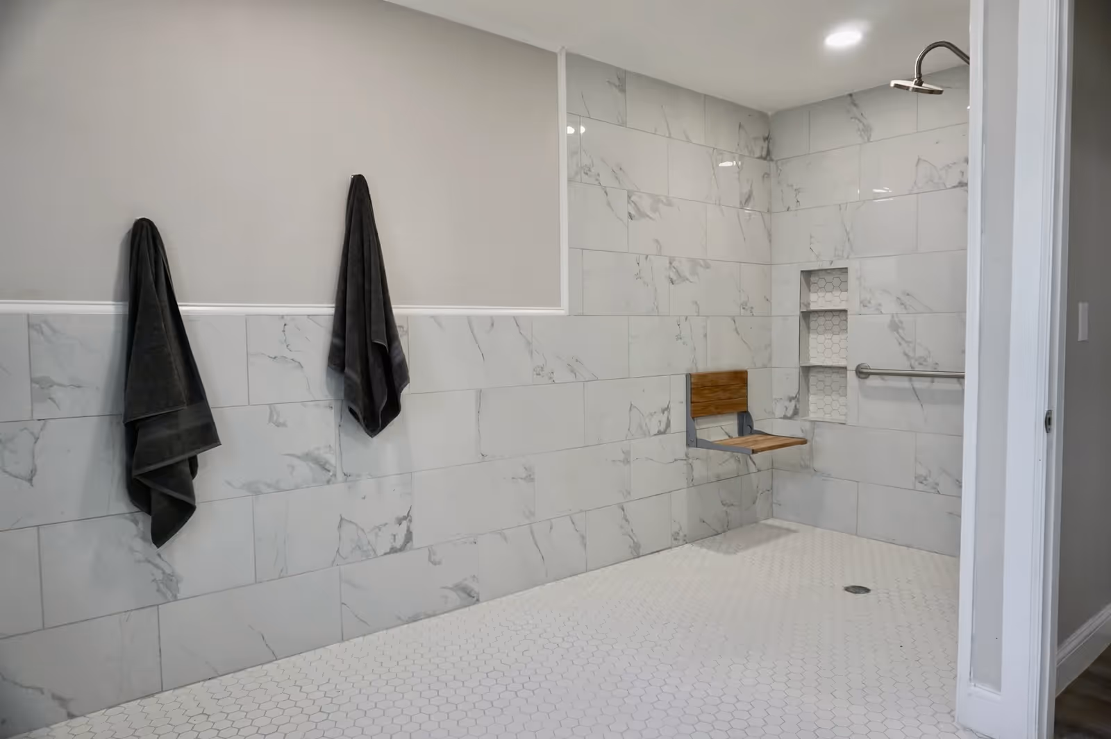 Accessible walk-in shower with marble tile walls, a fold-down wooden seat, grab bar, recessed shelf, and two dark towels hanging on the wall.