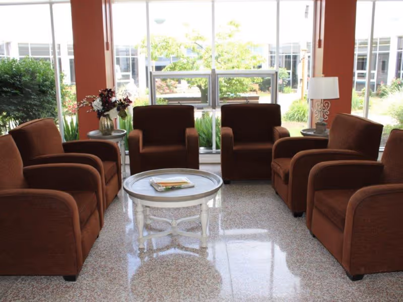 A seating area with six brown armchairs arranged around a round white coffee table in front of large windows overlooking greenery.