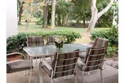 Outdoor patio area with a glass-top table and six cushioned chairs arranged around it. The patio overlooks a green garden with trees and shrubs in the background.