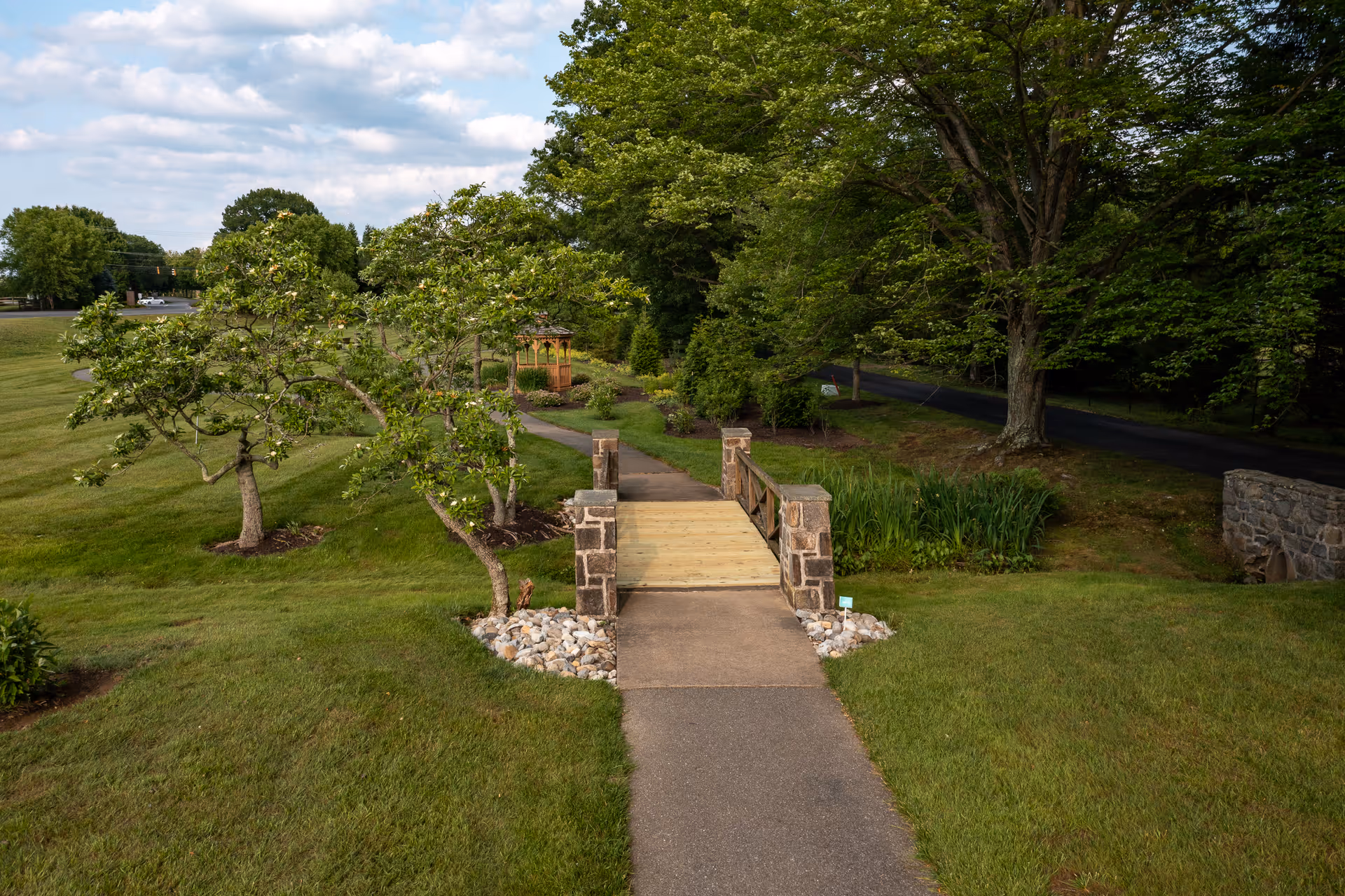 A paved path leads to a small wooden bridge and gazebo set in grassy, tree-lined landscaped grounds.