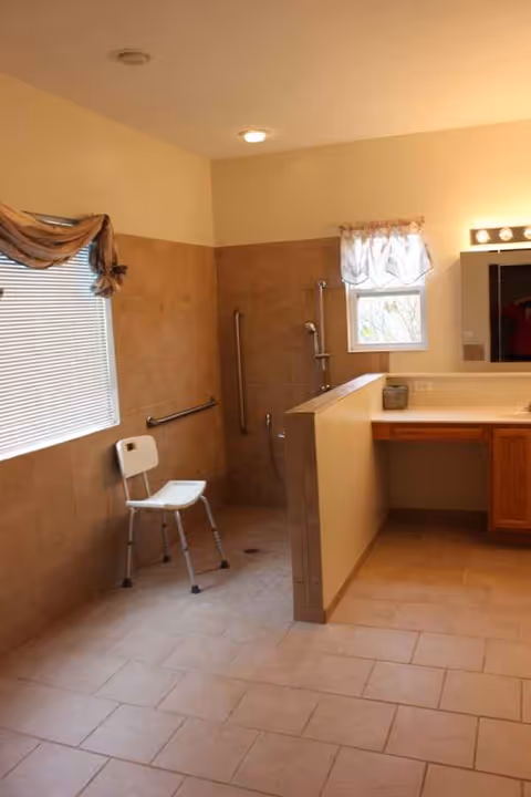 Accessible bathroom with beige tiles, a shower, chair, vanity, and window.