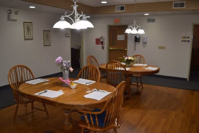 Interior dining area with two wooden tables, each set with placemats, utensils, and small flower arrangements. Wooden chairs surround the tables, and overhead lights hang above. The room has wooden flooring and white walls with minimal decorations. A door and a wall-mounted telephone are visible in the background.