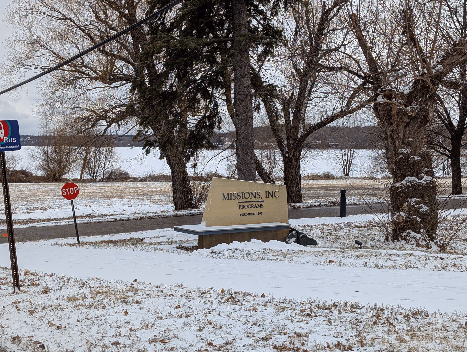 Outdoor scene showing a snow-covered ground with leafless trees and a beige stone sign that reads 'MISSIONS, INC PROGRAMS FOUNDED 1895'. There is a stop sign and a bus stop sign visible near a road in the background.