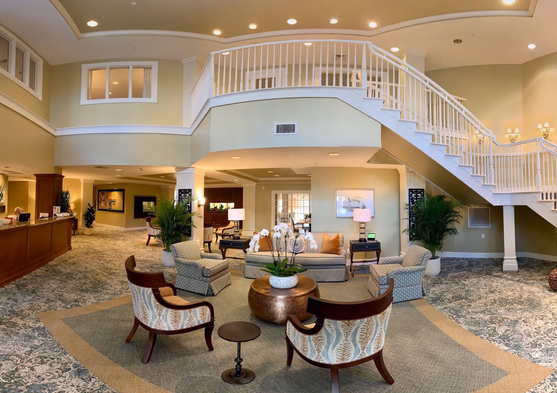 A spacious and well-lit senior living facility lobby with a seating area featuring patterned armchairs, a sofa, a round coffee table with a white orchid centerpiece, and side tables with lamps. The lobby has a high ceiling with recessed lighting, a staircase with white railings leading to an upper floor, and a reception desk on the left side. The walls are painted in a soft beige color, and there are large potted plants adding greenery to the space.